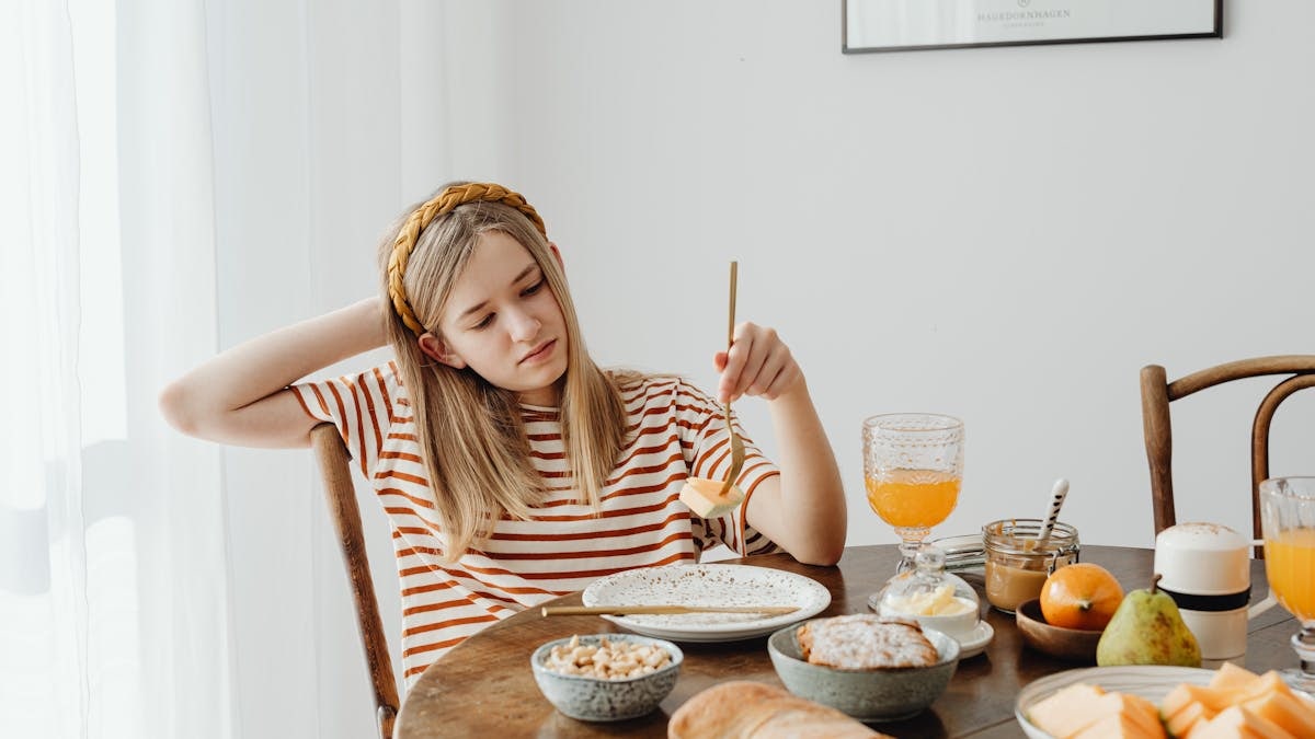 Teen girl in striped shirt at breakfast table with fruits, bread, and juice. Casual morning setting.